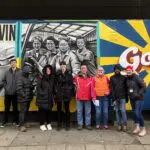 Group of people standing in front of a vibrant mural in Govan, featuring painted faces and the word "Meet me in Govan" with a sun emblem.