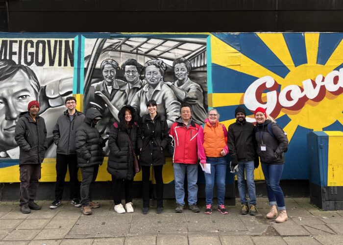 Group of people standing in front of a vibrant mural in Govan, featuring painted faces and the word "Meet me in Govan" with a sun emblem.