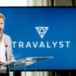 Man giving a presentation at a podium with the "Travalyst" logo on a screen behind him in a room with windows showing a daytime cityscape, attended by Prince Harry and representatives from travel and