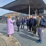 A tour guide speaking to a group of seven adults in front of the Welsh National Assembly building on a sunny day for the City of Imagination travel blog.