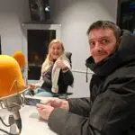 Two people in a recording studio, one giving a thumbs up. Both are seated and surrounded by microphones and electronic equipment, ready to record an episode of the "Invisible Cities Talks" podcast.