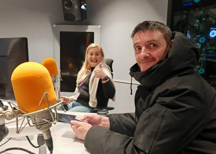 Two people in a recording studio, one giving a thumbs up. Both are seated and surrounded by microphones and electronic equipment, ready to record an episode of the "Invisible Cities Talks" podcast.