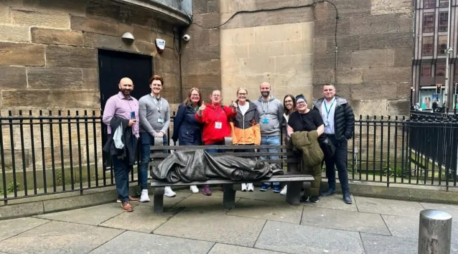 A group of people pose for a photo standing behind a bench with a statue of a person wrapped in a blanket lying on it, set against the backdrop of an alternative Glasgow stone building.