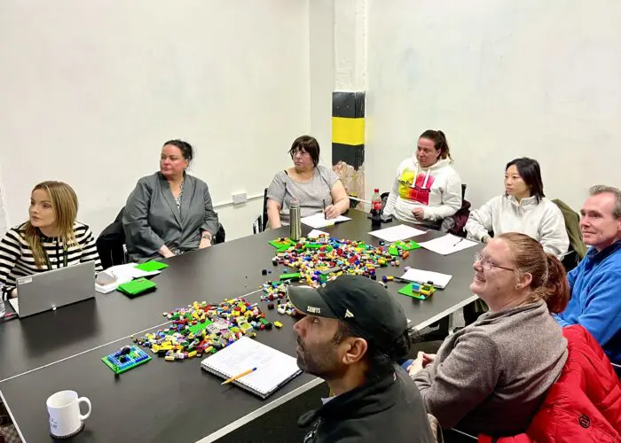 A group of people sit around a table with Lego bricks in the center, attentively watching something off-camera. Their training ethos is evident through the notebooks, pens, and cups on the table, indicating a methodical approach to their activity.