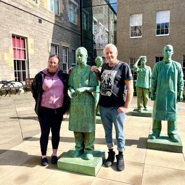 Two people stand outdoors in Edinburgh beside life-sized green statues of medical innovators, with more statues and city buildings in the background.