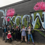 A group of six people poses in front of a mural adorned with "Govan" and pink flowers on a brick wall, reminiscent of the vibrant street art near SWG3 Glasgow.