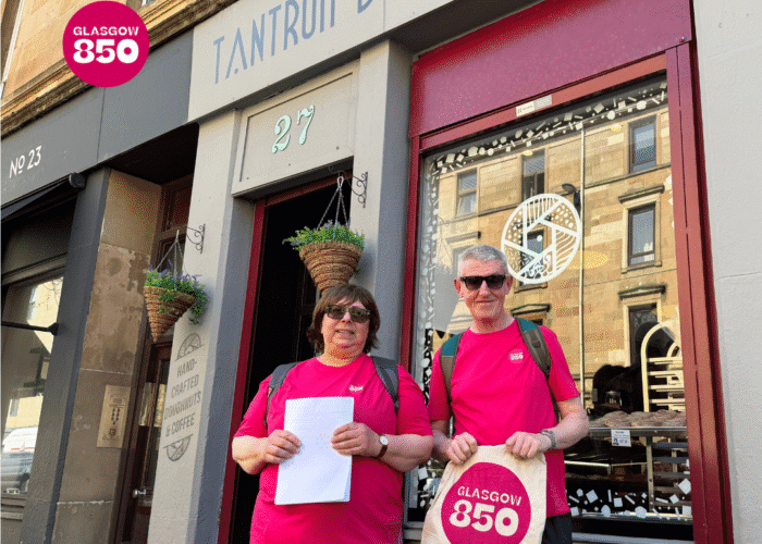 Two people in matching pink shirts stand outside Tantrum Doughnuts, embracing Glasgow hospitality with a Glasgow 850 tote bag and paper. The shop features hanging plants and a sign above the door.