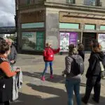A tour guide from Invisible Cities in a red jacket speaks to a group of people outside a Glasgow building with informational posters and boarded-up windows.
