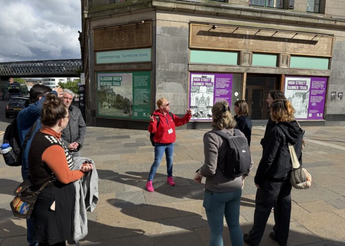 A tour guide from Invisible Cities in a red jacket speaks to a group of people outside a Glasgow building with informational posters and boarded-up windows.