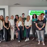 A group of thirteen women stand together in a conference room, smiling for a group photo in front of a screen that reads "Women Supporting Women in Manchester" at one of the city's dynamic B2B events.