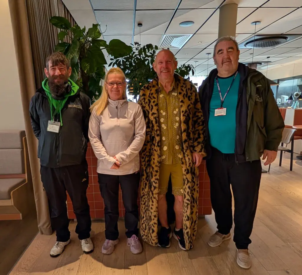 Four adults stand indoors in a row, facing the camera. One person wears a leopard print coat. Plants and tables decorate the background, creating a welcoming Liverpool vibe—perhaps inspired by Invisible Cities tours.